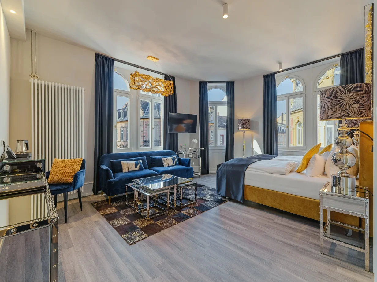 Heller Superior Room mit hohen Fenstern, Doppelbett, farbenfroher Einrichtung in gold, weiß und blau sowie einem Sitzbereich im BS Boutique Hotel Heidelberg.